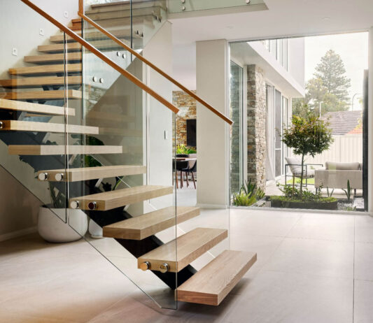Floating timber staircase with glass balustrading, timber handrail and double-height glazing in a custom home.