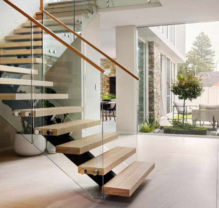 Floating timber staircase with glass balustrading, timber handrail and double-height glazing in a custom home.