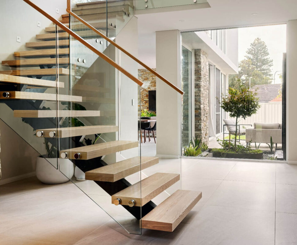Floating timber staircase with glass balustrading, timber handrail and double-height glazing in a custom home.