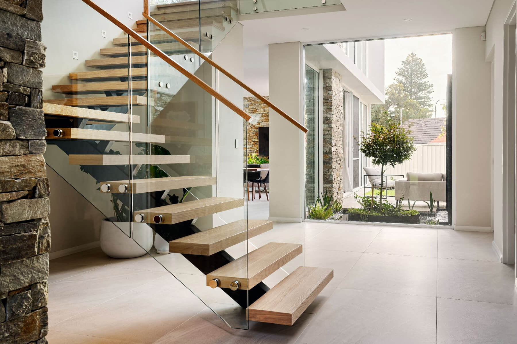 Floating timber staircase with glass balustrading, timber handrail and double-height glazing in a custom home.