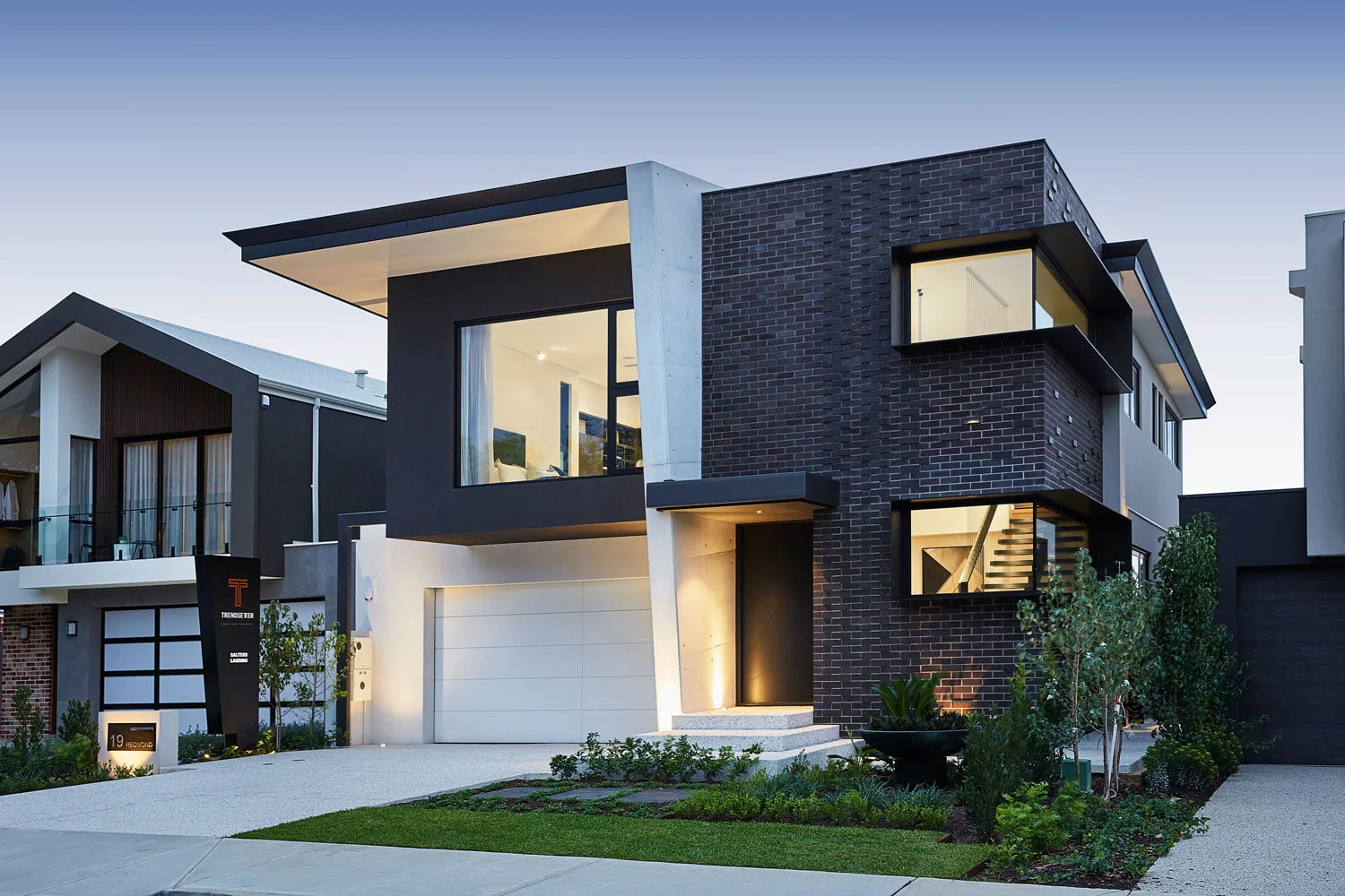Contemporary two-storey custom home featuring dark brickwork, angular rooflines and illuminated interiors at dusk.