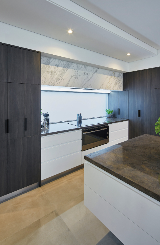 Minimalist kitchen featuring dark handleless cabinetry, stone benchtops and large windows.
