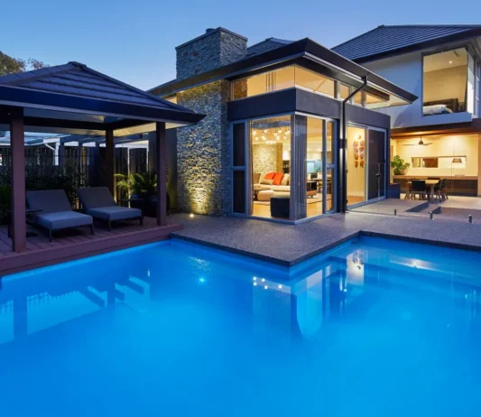 Custom home swimming pool and alfresco area featuring timber decking, expansive glazing and outdoor lighting at dusk.