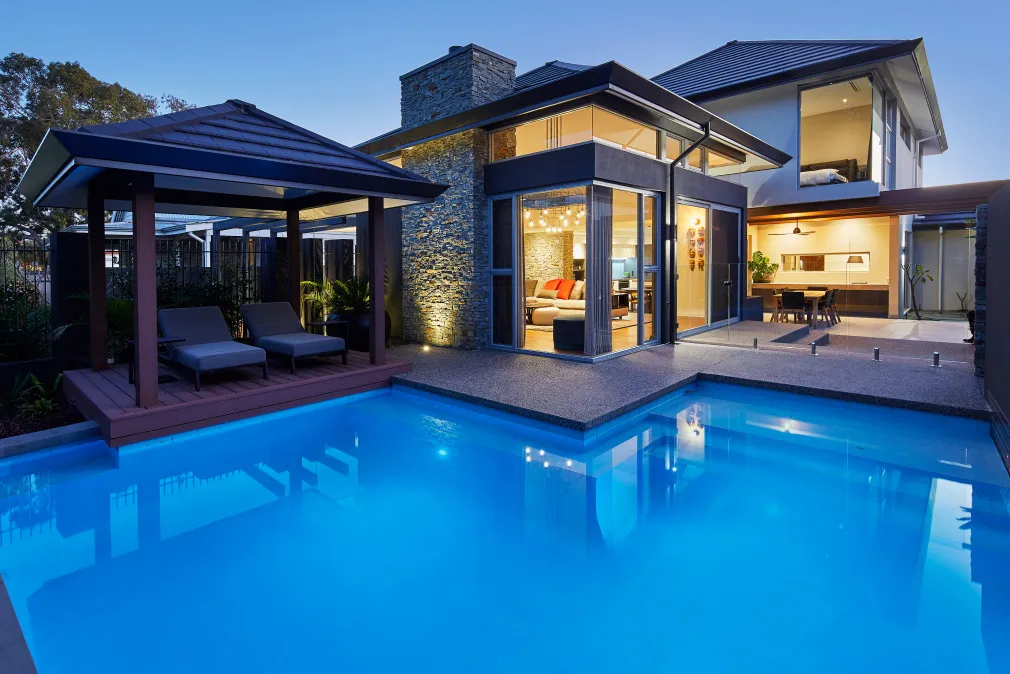 Custom home swimming pool and alfresco area featuring timber decking, expansive glazing and outdoor lighting at dusk.