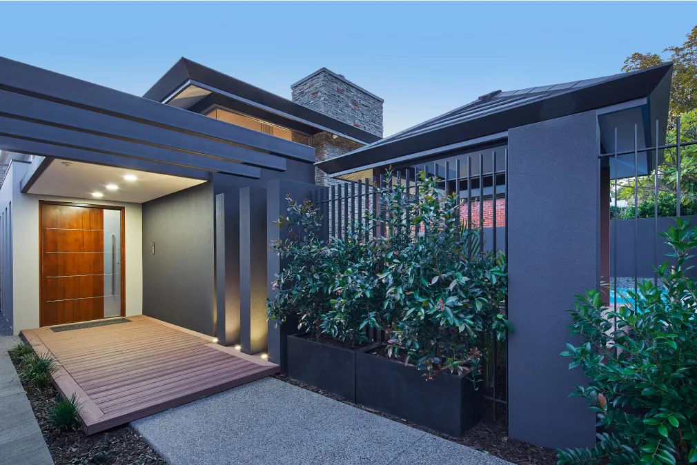 Exterior walkway alongside a custom home featuring dark cladding, architectural lighting and landscaped greenery.