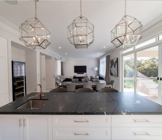 Open-plan kitchen featuring a large island bench, decorative pendant lights and expansive glazing.