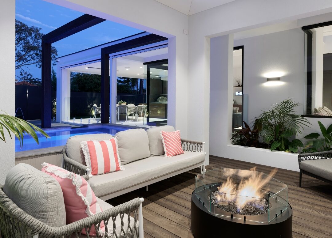 Covered outdoor living area featuring a central fire pit, woven lounge furniture, timber decking and views of the swimming pool at dusk.