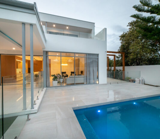 Custom home swimming pool surrounded by paved outdoor living areas and large windows connecting to the interior.