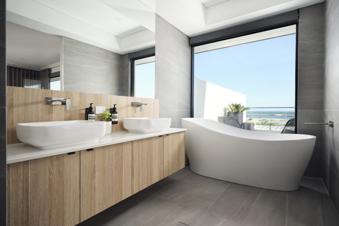 Coastal custom home kitchen featuring light-coloured cabinetry, stone benchtops, freestanding bath and large windows overlooking the ocean.